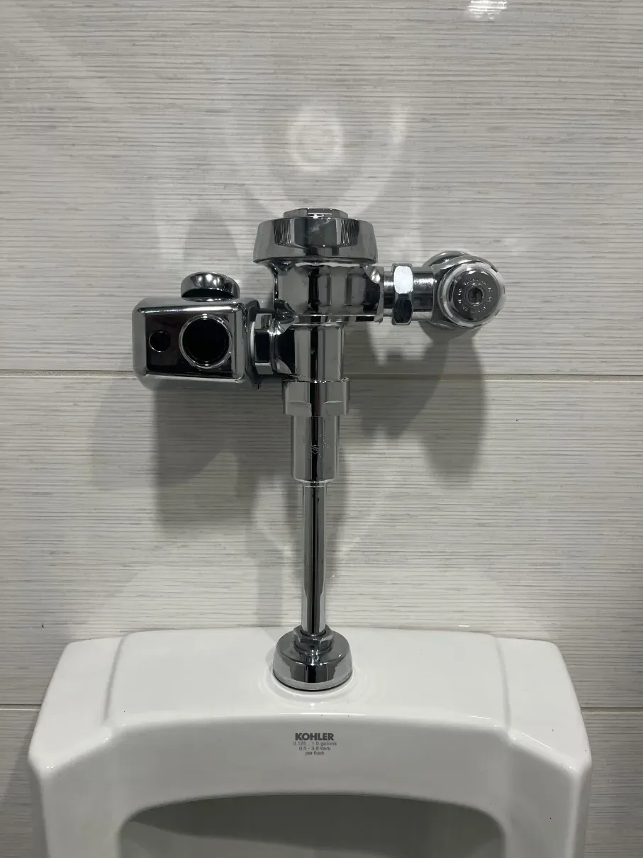 Chrome urinal flush valve on a white wall-mounted urinal against a light textured wall