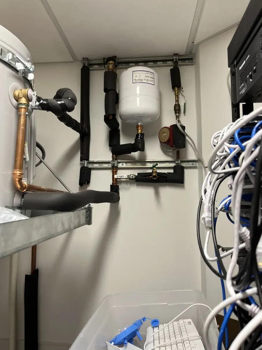 Utility closet with pipes, a white tank, and cables around network equipment.