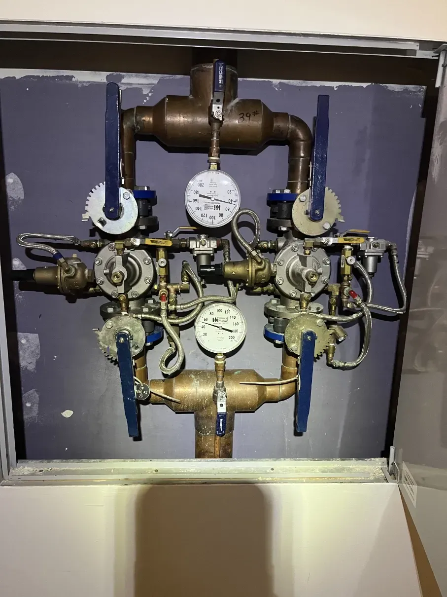 Open utility valve manifold with brass pipes, blue handles, and pressure gauges inside a wall cavity