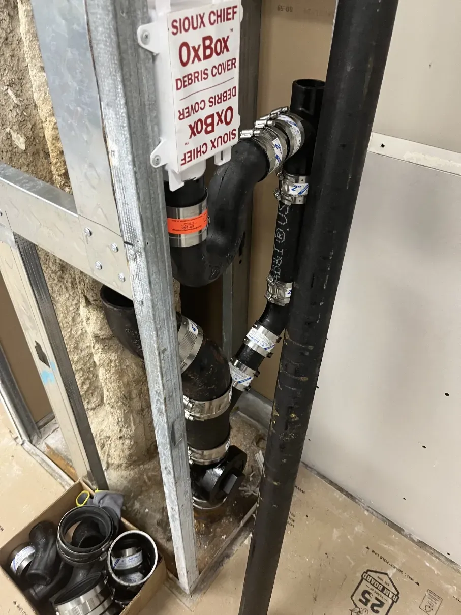Plumbing pipes and fittings installed in a wall cavity beside a white panel, with a metal stud frame.