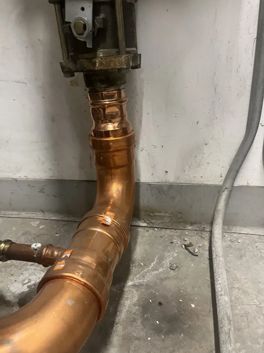 Copper plumbing pipe connected to a wall-mounted valve above a concrete floor, with signs of leakage.