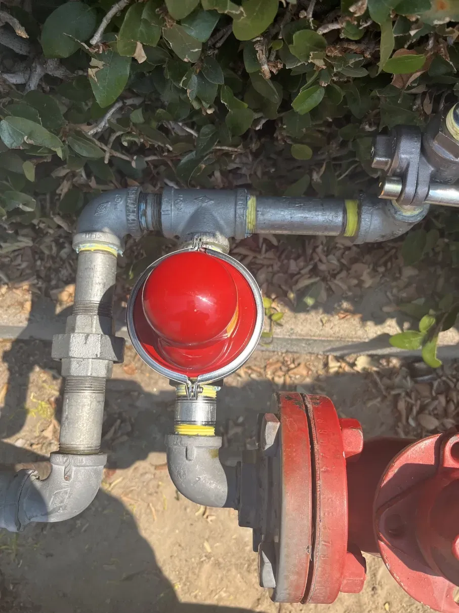 Red sprinkler/valve assembly with metal pipes and a red hydrant connection in a dirt area near shrubs