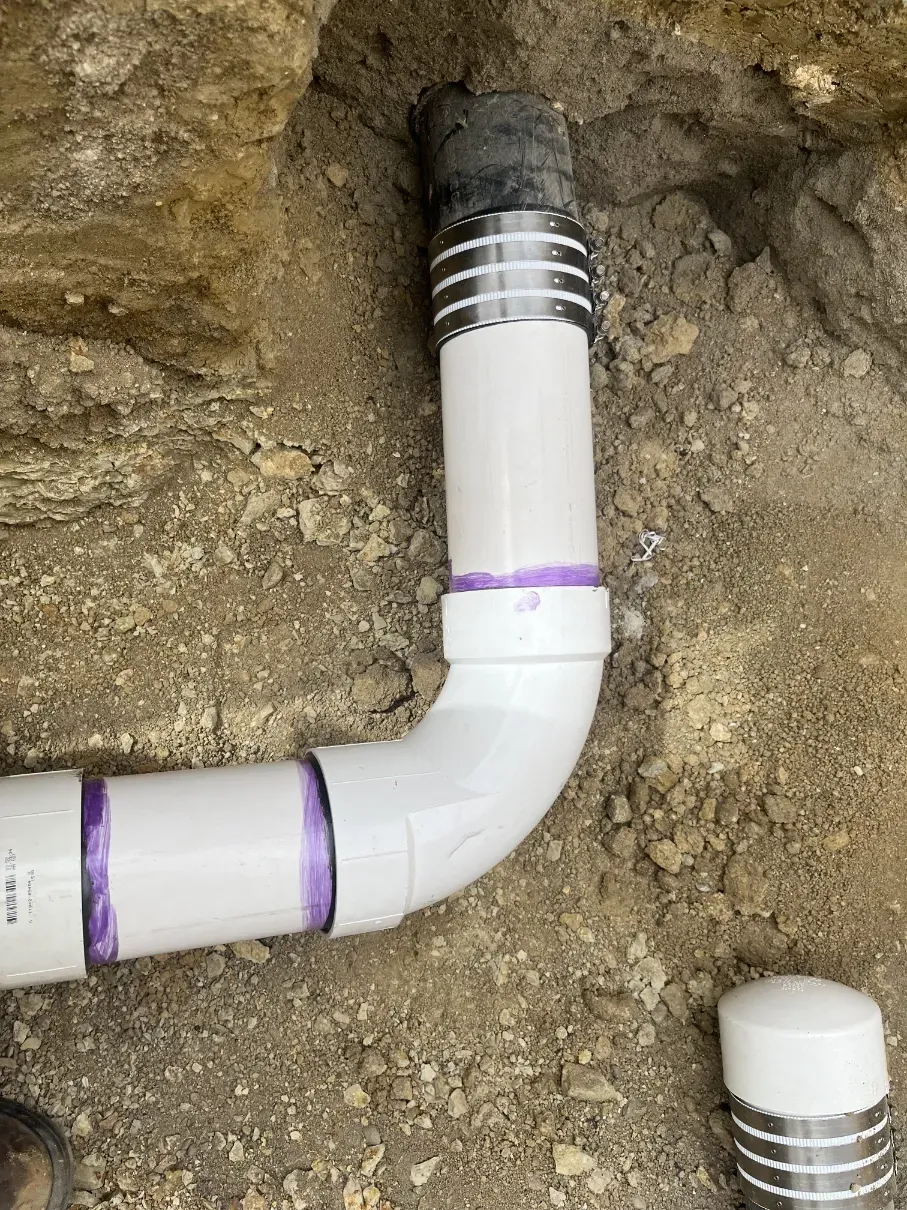 White PVC pipe elbow in dirt trench with black fitting and purple markings; pipe cap nearby