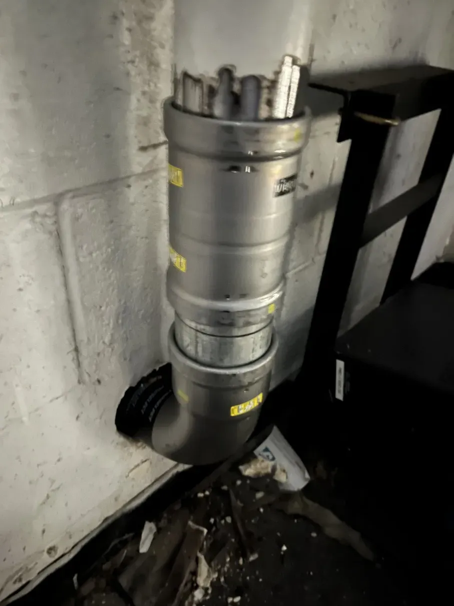 Gray pipe or vent duct against a concrete wall, with dark debris on the floor in a utility space
