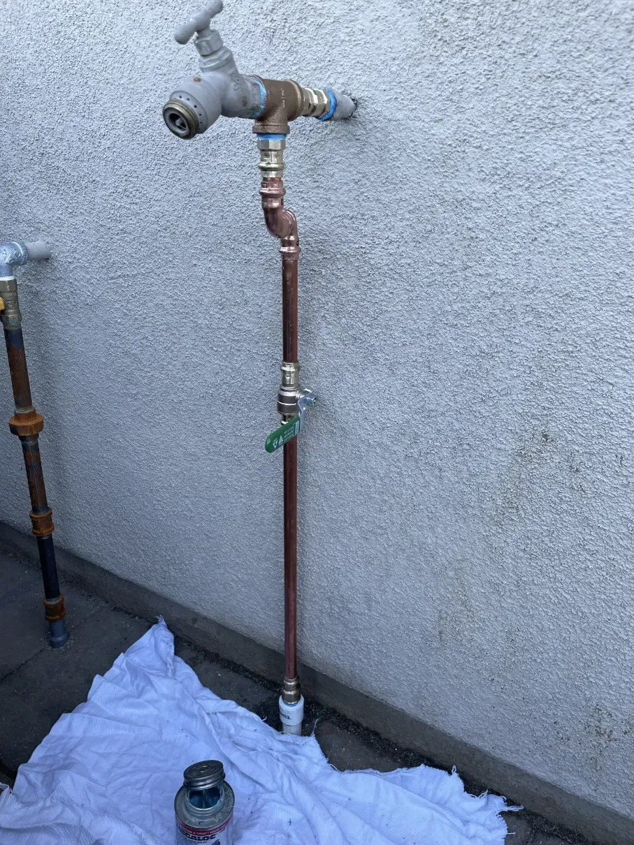 Outdoor wall-mounted water spigot with a copper pipe and a bucket on the ground.