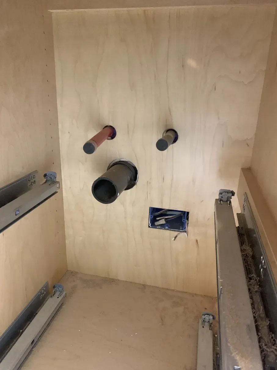 Wooden utility cabinet interior with plumbing pipes and metal drawer rails