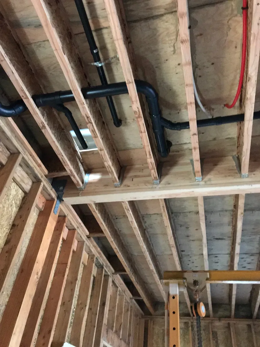 Exposed wooden ceiling joists and wall studs in an unfinished room with visible pipes and wiring
