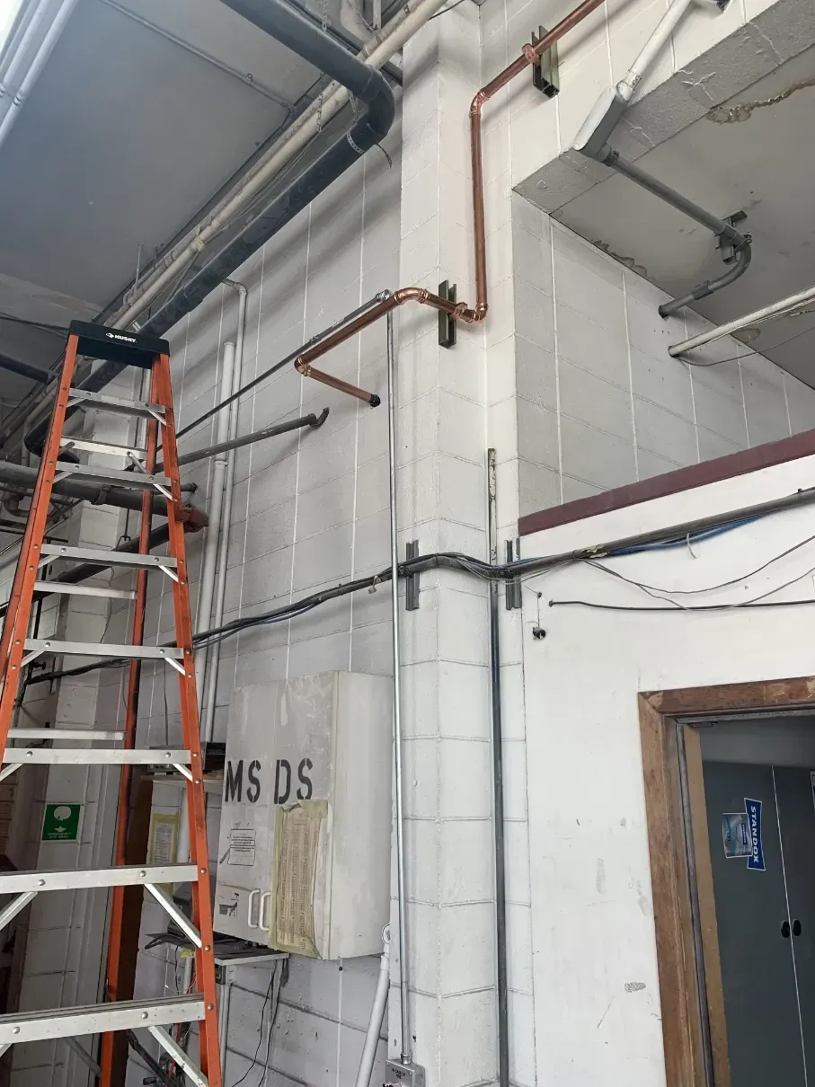 Orange ladder beside white industrial wall with exposed pipes and wiring in a construction area