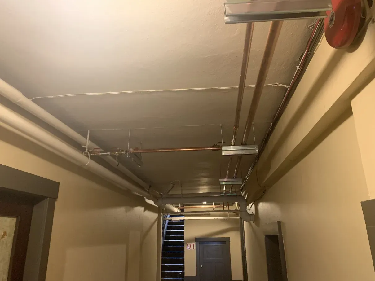Narrow hallway with overhead pipes and ceiling fixtures, leading to a door at the far end.