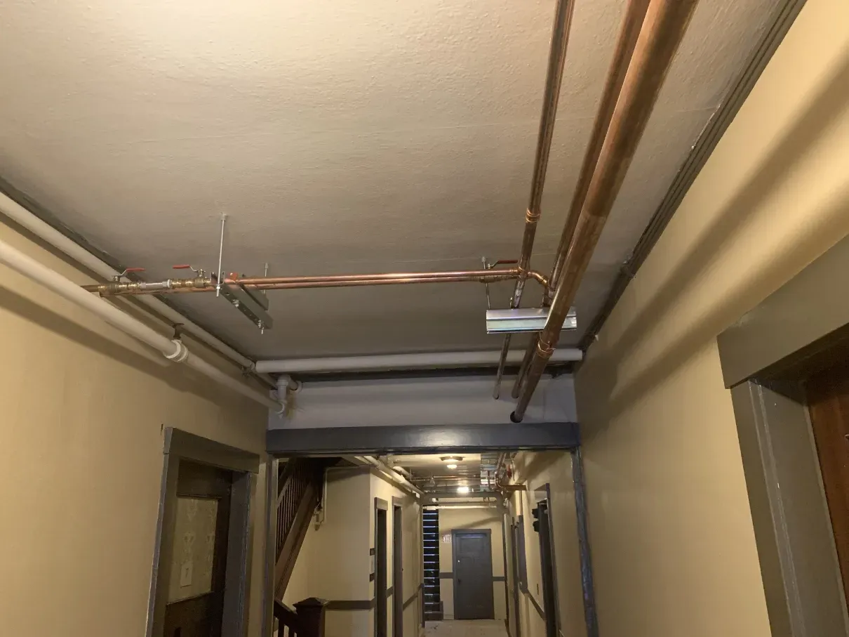 Narrow indoor hallway with exposed pipes and ceiling fixtures, lit by fluorescent lights