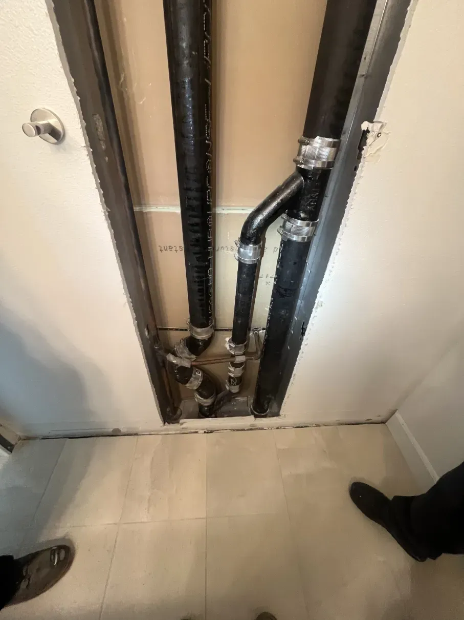 Open utility wall cavity with exposed black pipes and white floor tiles, viewed from above