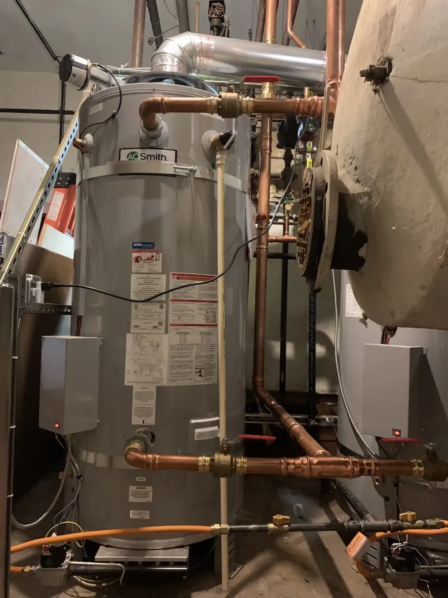 Water heater and copper pipes in a utility room, with a large white tank on the right.