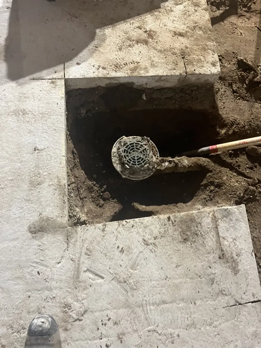 Concrete slab with a square hole exposing a pipe, with a tool inserted into the opening.