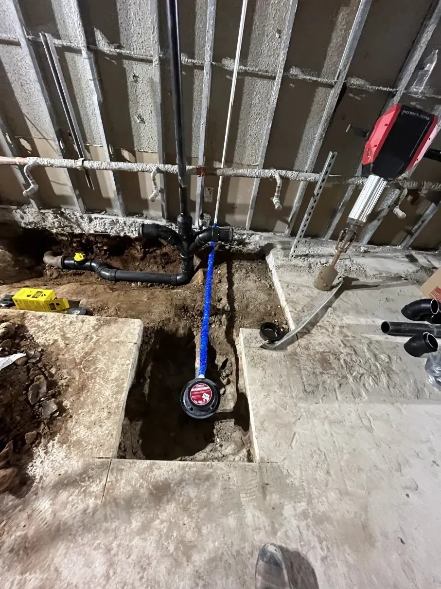 Exposed pipes and plumbing lines in a concrete floor trench beneath a metal wall frame.