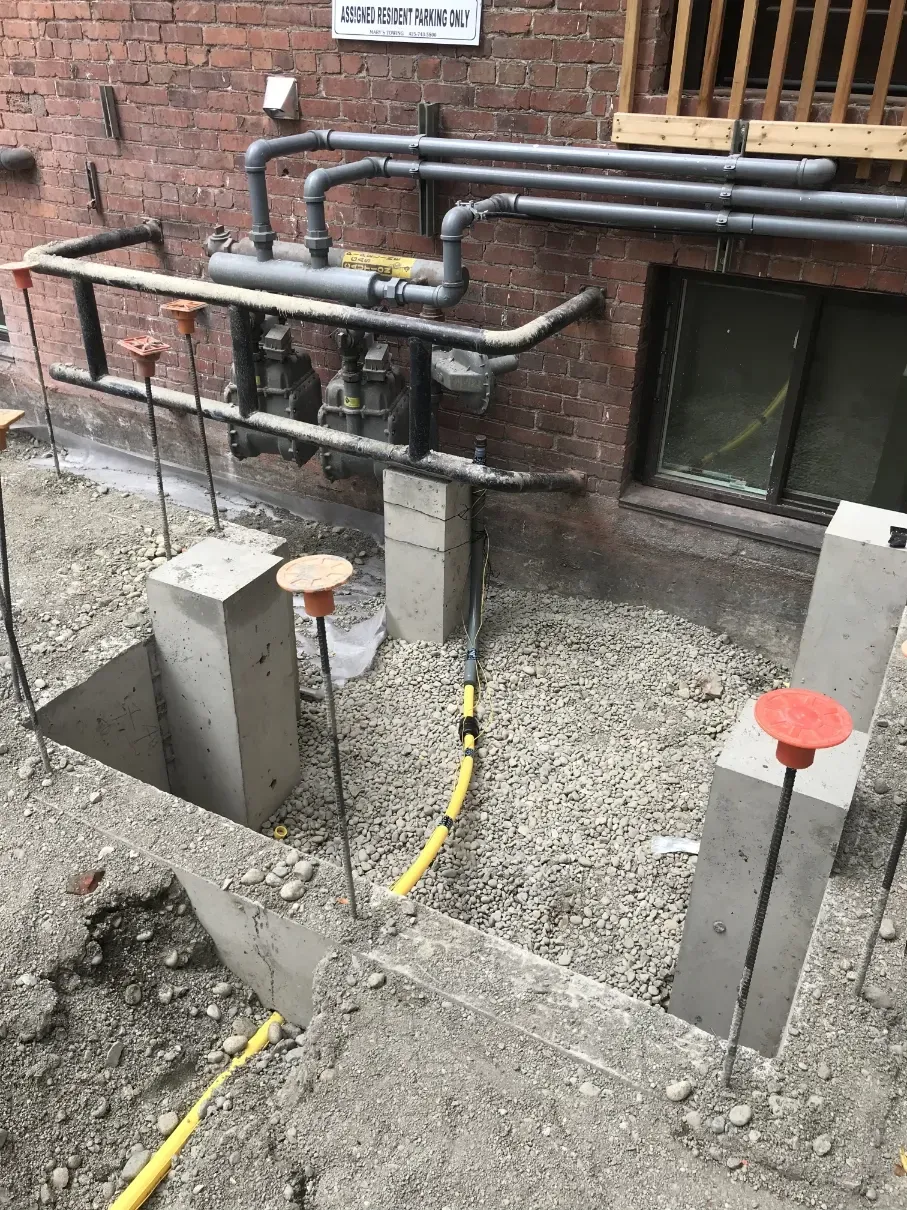 Backyard utility trench with exposed pipes, concrete footings, and gravel beside a brick wall.