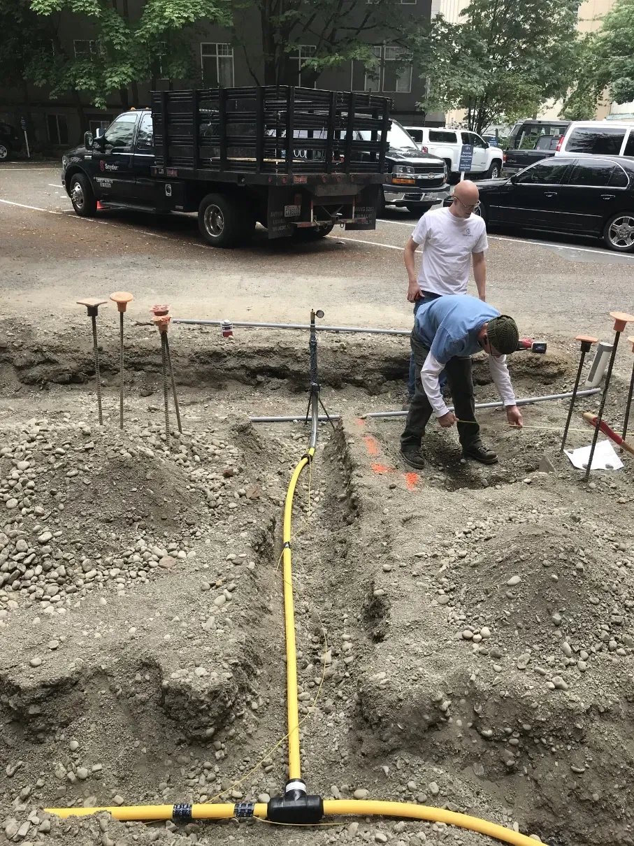 Workers at a road excavation site with a yellow cable and parked trucks nearby