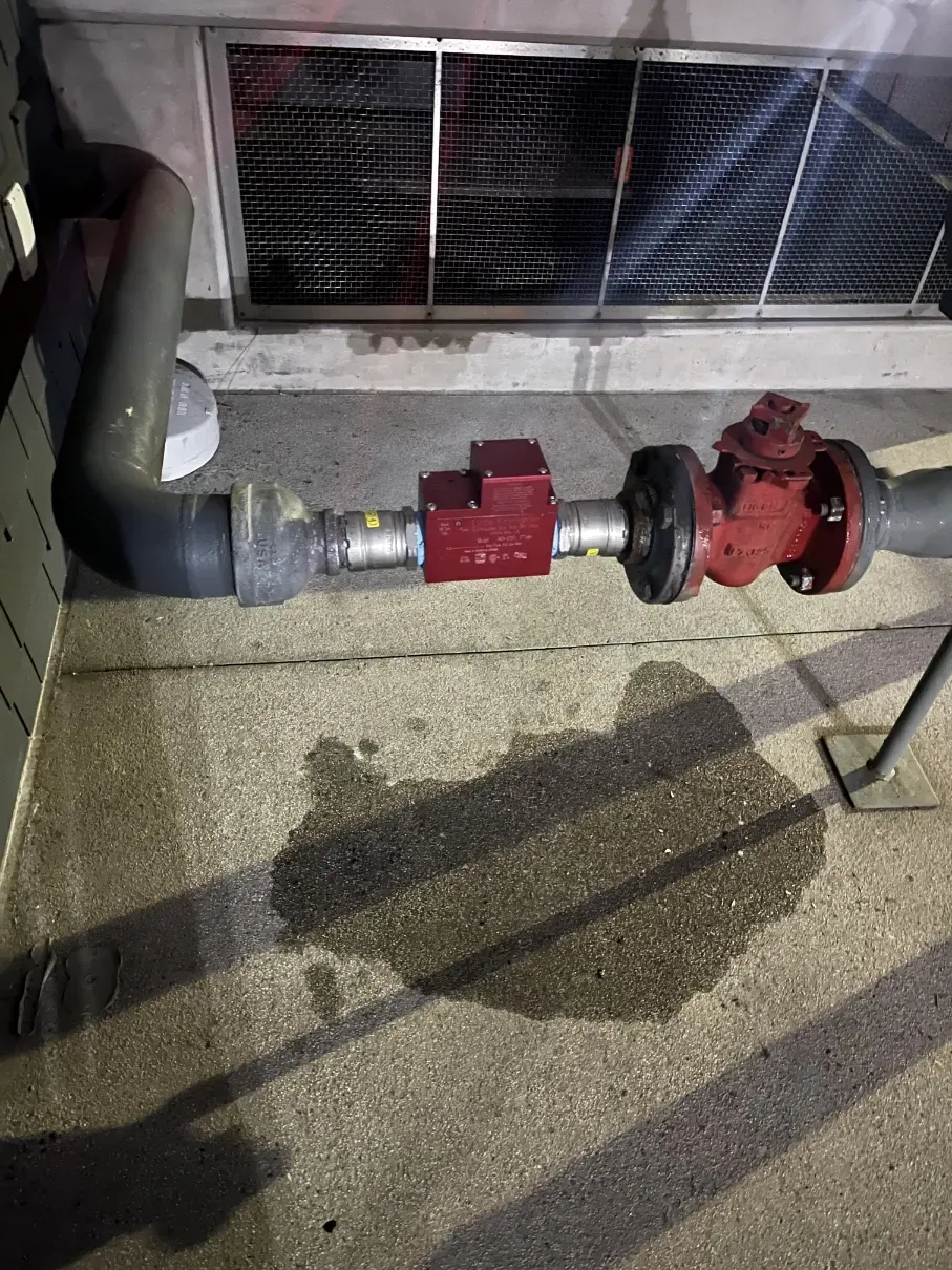 Red and gray industrial pipe assembly with valve on a concrete floor beneath a vent