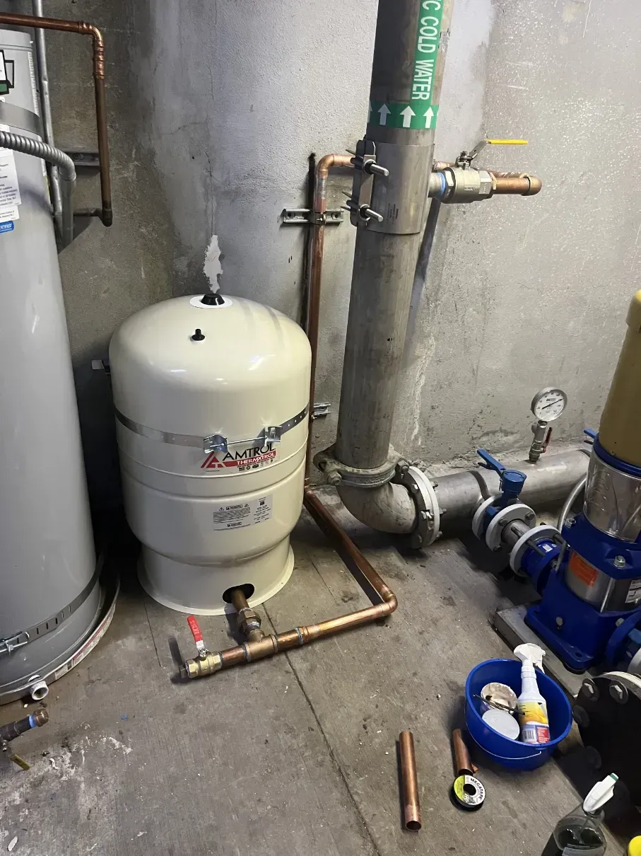 Basement utility area with white pressure tank, pipes, valves, and plumbing tools on the floor.