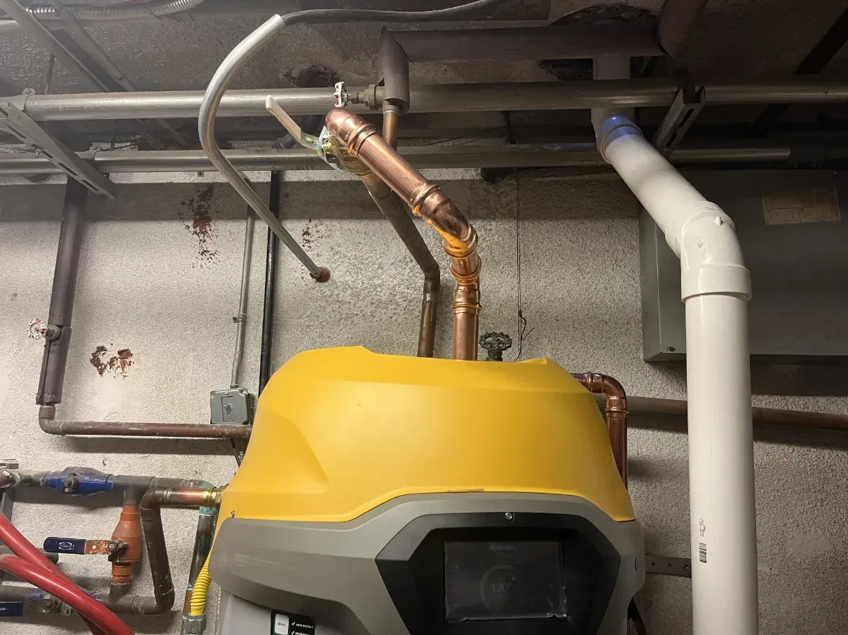 Yellow industrial machine below exposed copper and white pipes in a utility room