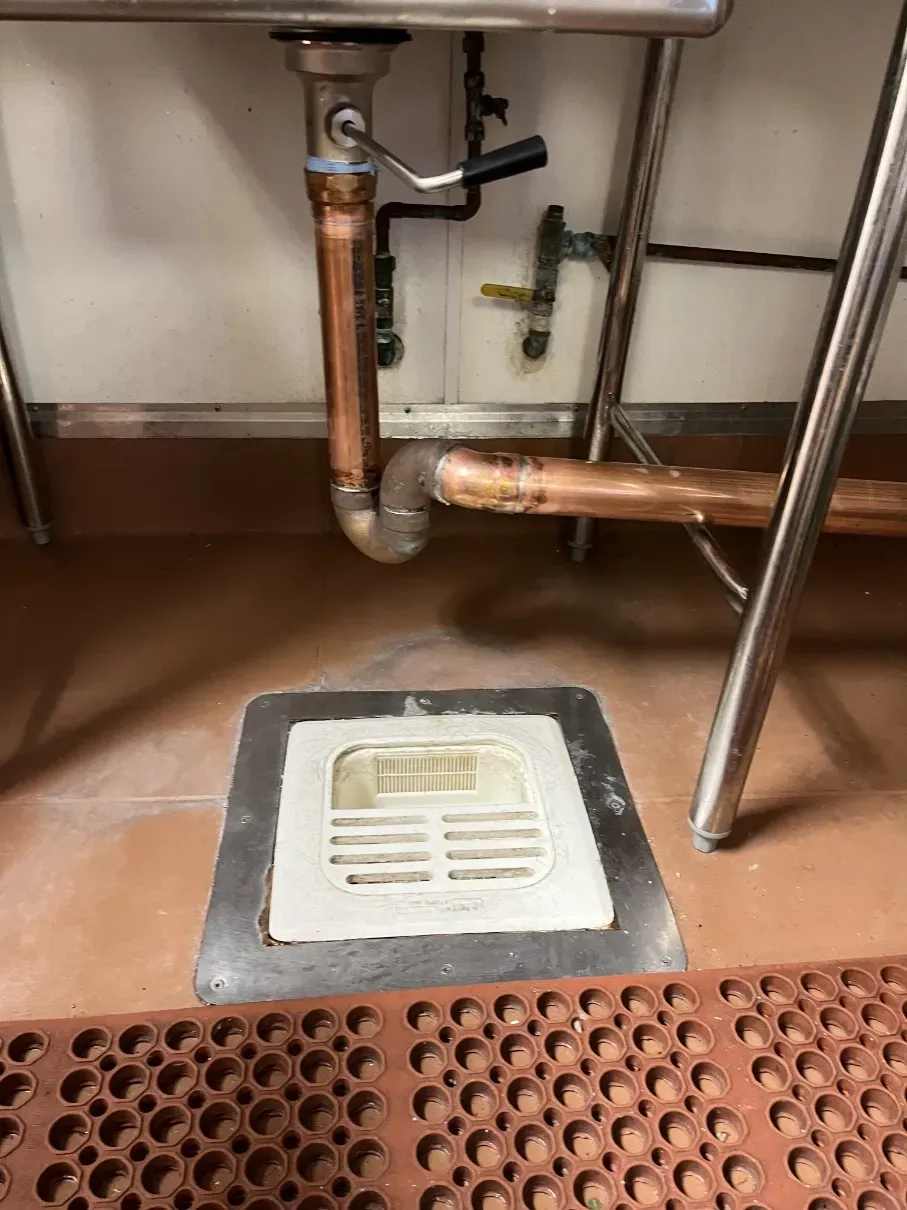 Floor drain beneath exposed copper plumbing in a bathroom or utility area.