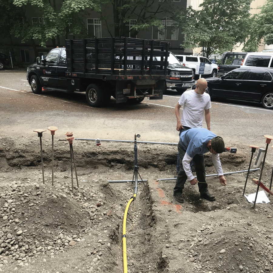 Workers digging a trench beside a black truck in a city street construction site.