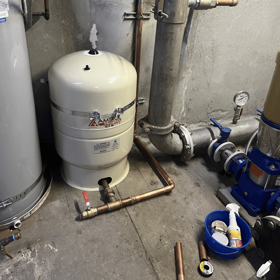 White industrial tank and blue pump equipment in a utility room with pipes and gauges