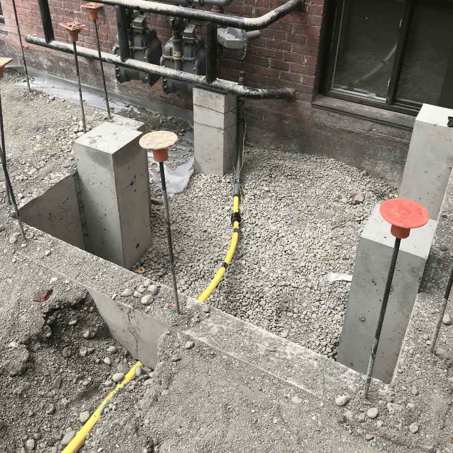 Concrete footings with rebar and yellow conduit near a brick wall, surrounded by gravel, ready for construction work.