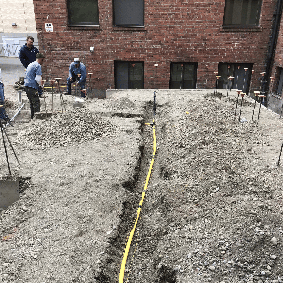 Workers on a construction site lay a yellow cable in a trench near a brick building, surrounded by gravel and metal rods.