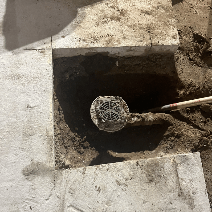 A partially dug hole in concrete reveals a drain cover, with a shovel nearby, highlighting ongoing construction work.