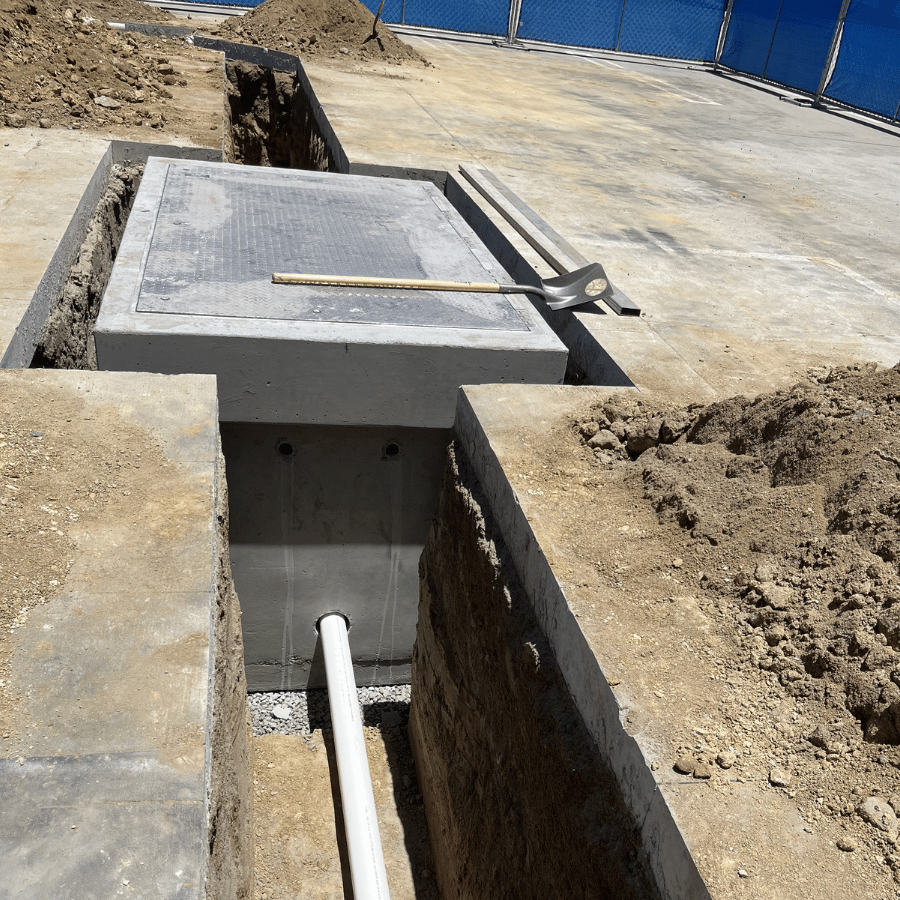 Construction site showing a concrete trench with a pipe, shovel, and measuring stick, surrounded by dirt and blue fencing.