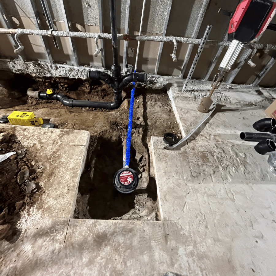 Construction site with exposed pipes, a trench in concrete floor, and tools scattered around, indicating ongoing plumbing work.