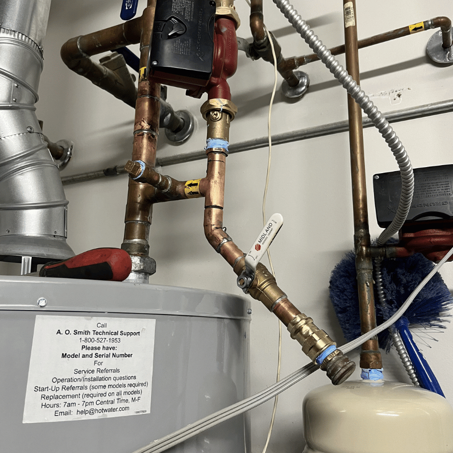 Copper pipes and valves connect to a water heater. A label with technical support details is visible on the heater's surface.
