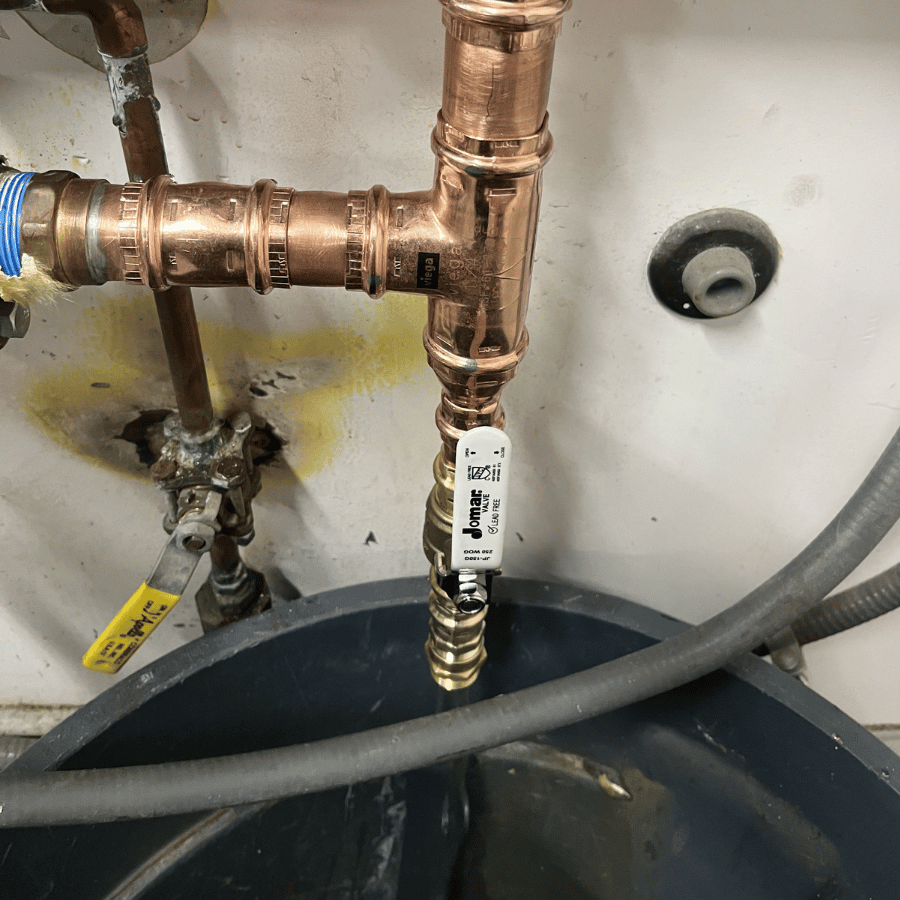 Copper pipes with a Jomar valve are connected above a black container, with hoses and a yellow valve nearby on a white wall.