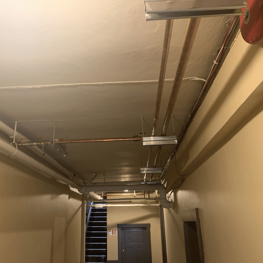 Dimly lit hallway with exposed pipes on the ceiling, leading to a door and an exit sign near a staircase.