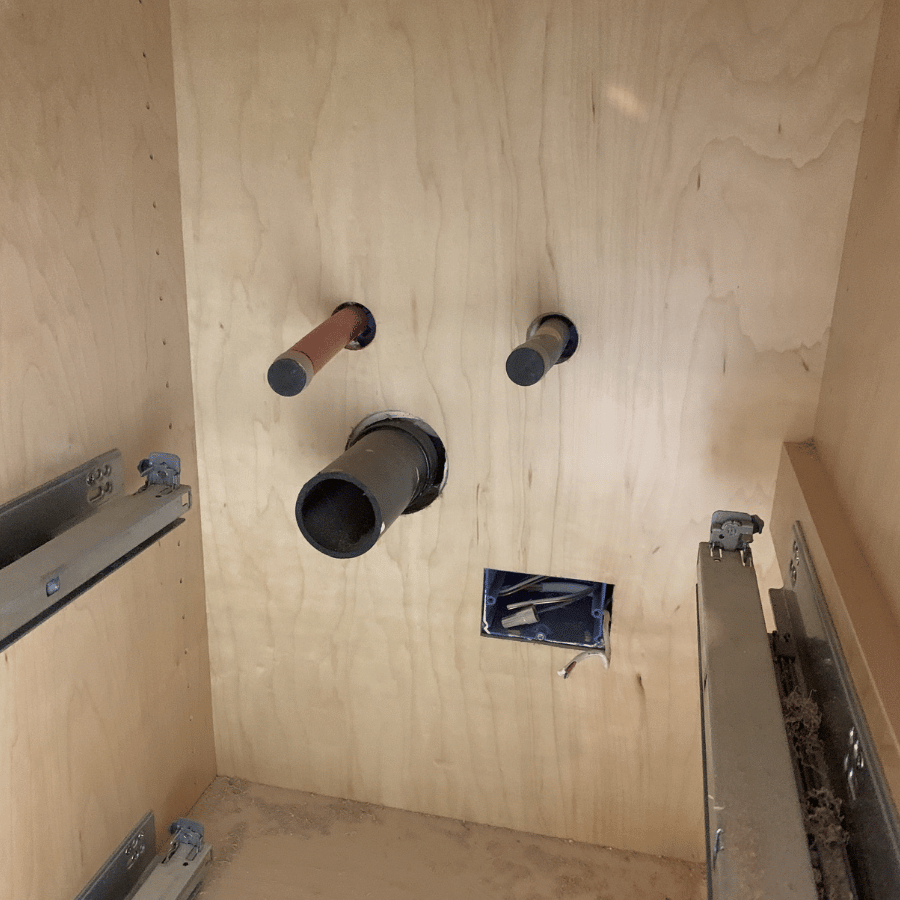 Wooden cabinet interior showing plumbing pipes and an electrical box with wires, alongside drawer slides on both sides.