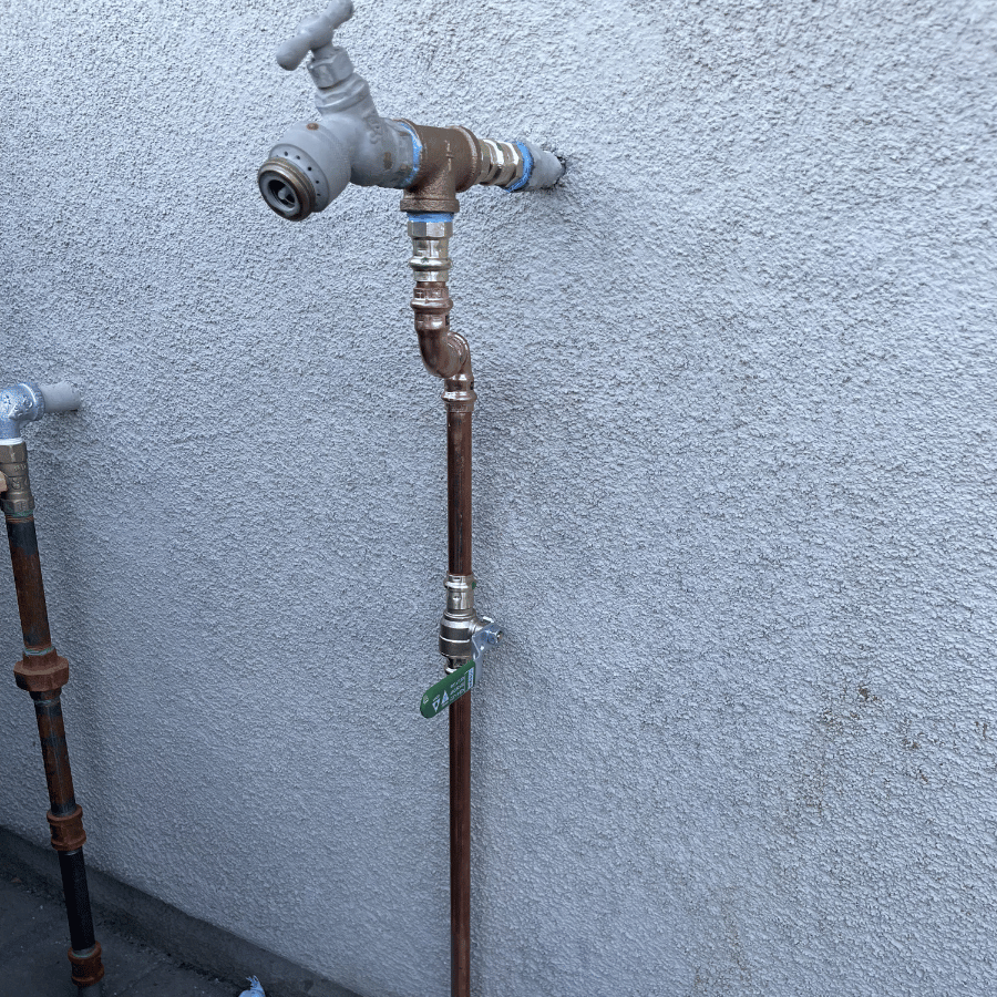 Outdoor faucet with copper piping against a textured wall, featuring a green valve tag and additional pipe on the left.