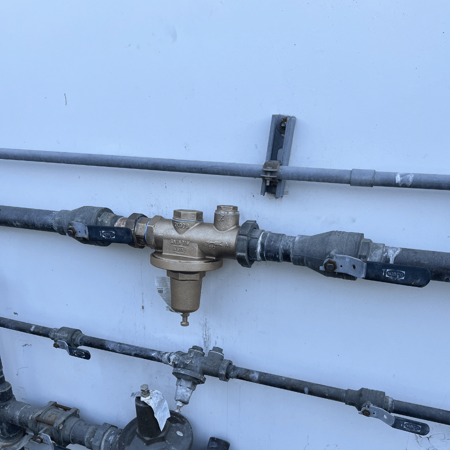 Brass valve with gauges and pipes on a white wall, part of a plumbing system, showing connections and fittings.