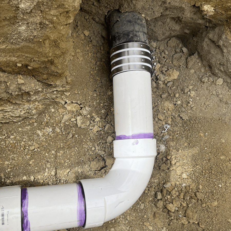 A white PVC pipe with purple sealant joins a black pipe underground, surrounded by dirt, indicating plumbing work.
