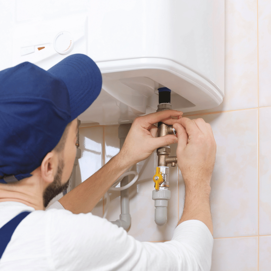 A plumber in a blue cap repairs a wall-mounted water heater, adjusting pipes and valves in a tiled room.