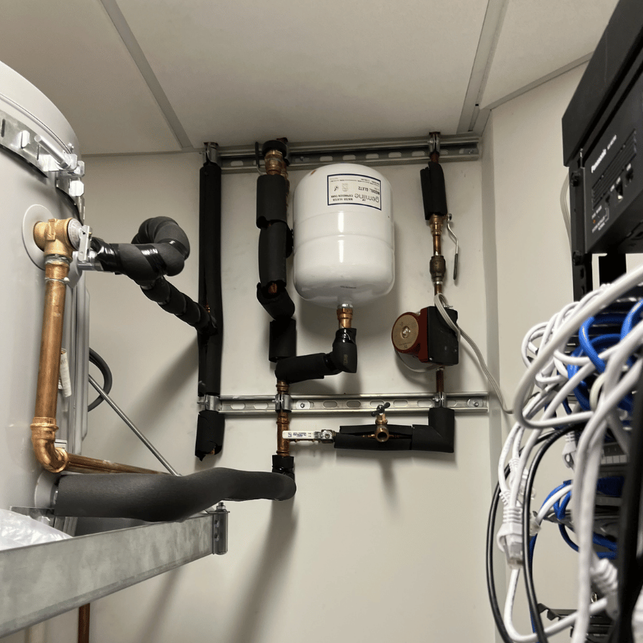 Pipes and cables in a utility room, featuring a white expansion tank and insulated pipes, organized neatly on the wall.
