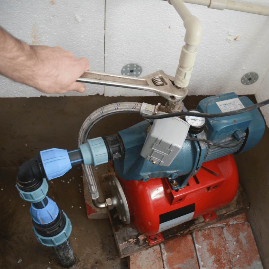 A hand uses a wrench to adjust a pipe connection on a blue and red water pump in a basement, ensuring secure installation.