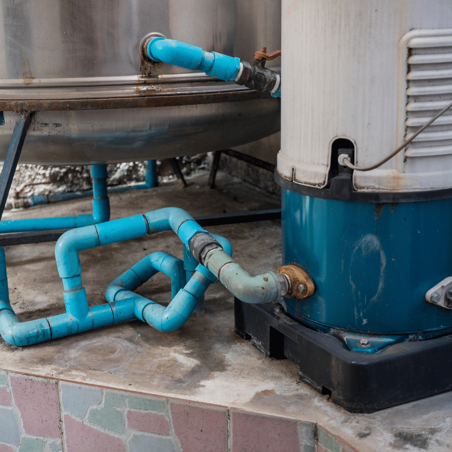 Blue pipes connect a large metal tank to a cylindrical water heater on a tiled floor, showing a plumbing setup.