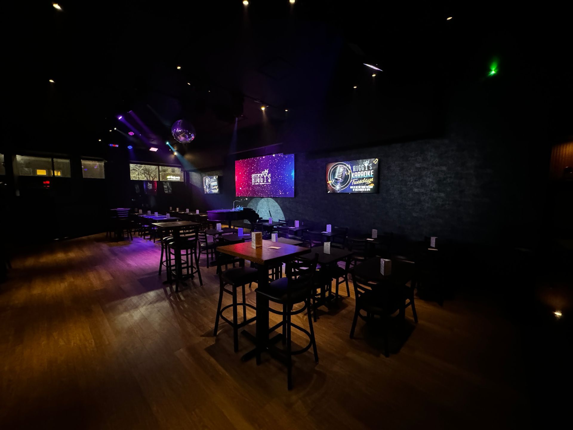 Interior of a dimly lit bar with tables, chairs, and several screens; wooden floors, disco ball.
