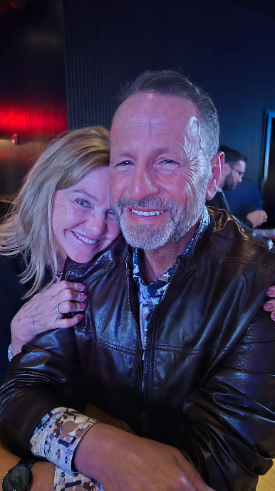 Woman embracing a man; both smiling. Man wearing a brown leather jacket. Dark indoor setting.