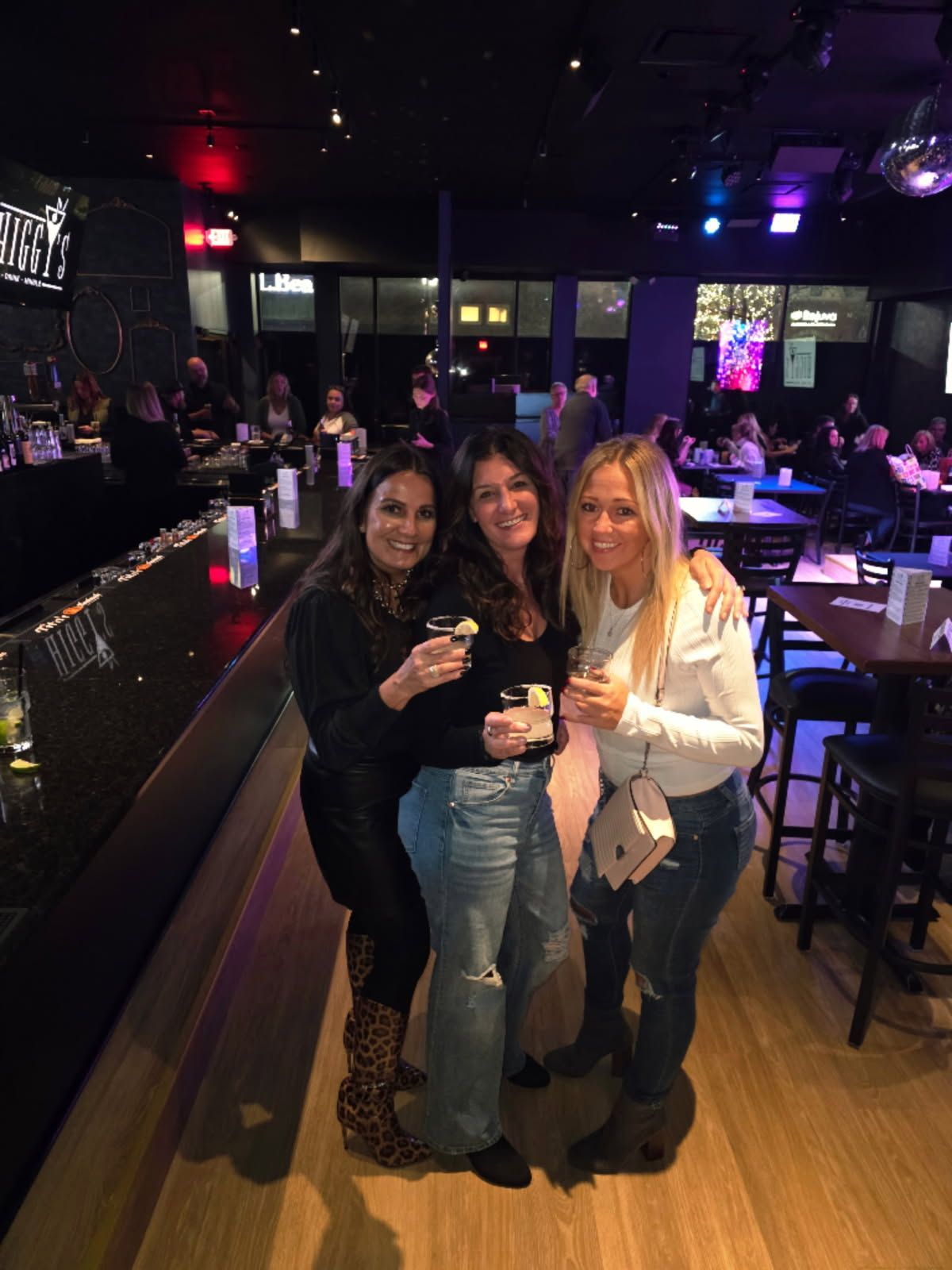 Three women at a bar, holding drinks. One wears leopard print boots, smiling. Dark interior, people in background.