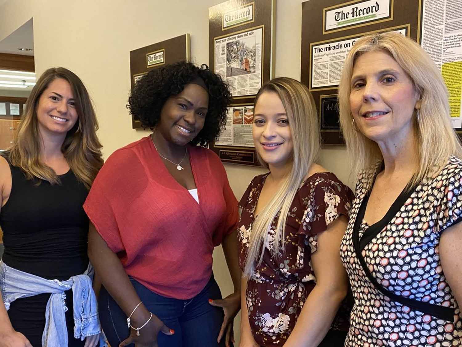 Four women smiling, posing for a photo. They stand against a wall with framed newspaper clippings.