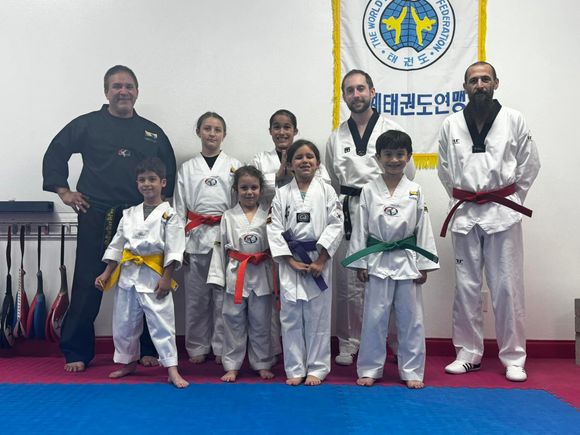 A diverse group in martial arts uniforms stands together in a taekwondo studio, posing in front of a banner.