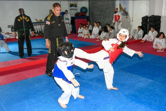 Two taekwondo students in gear practice a kick in a studio, with their instructor and classmates watching nearby.