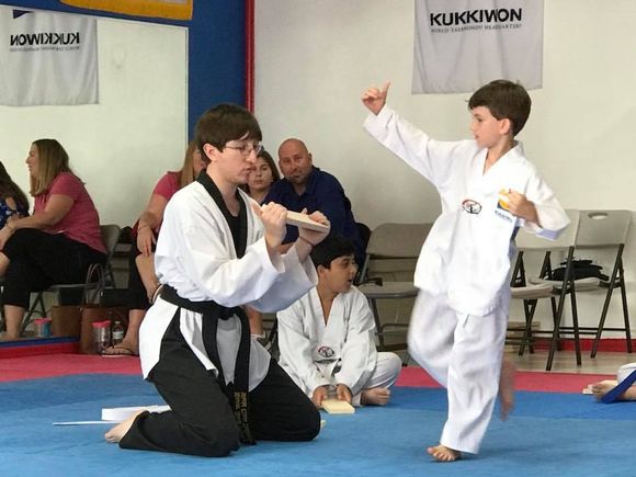 An instructor in a black belt uniform holds a wooden board for a student in a white uniform to practice a kick in a dojo.