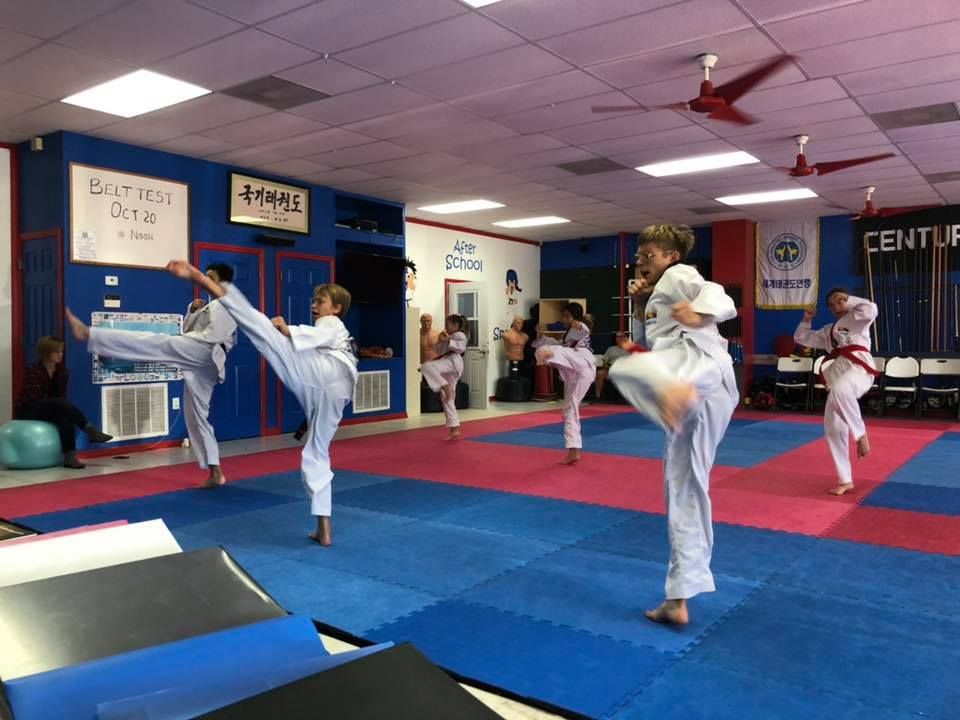 Students in white karate uniforms practice high kicks on a blue and red mat in a martial arts studio.